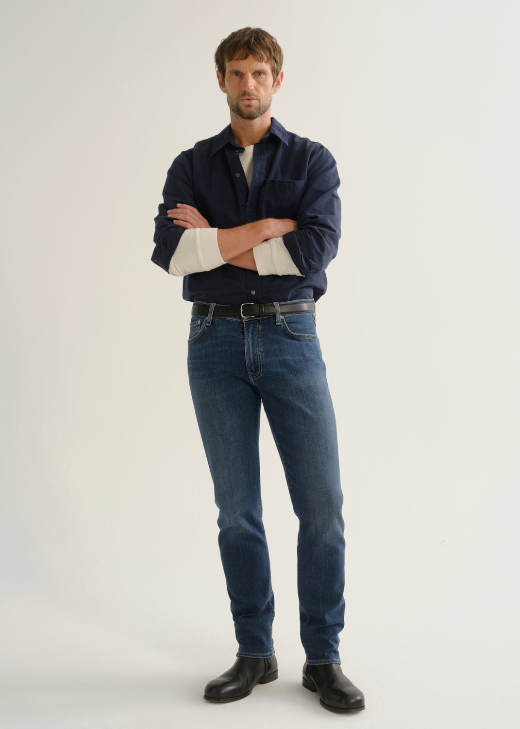 Person wearing medium blue straight leg jeans with a navy long sleeve button-up shirt and black shoes, front full body view on a light grey background.