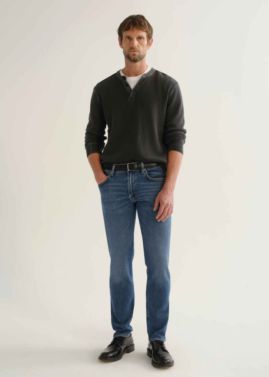 Person wearing medium blue straight leg jeans with a black long sleeve shirt and black shoes, front full body view on a light grey background.