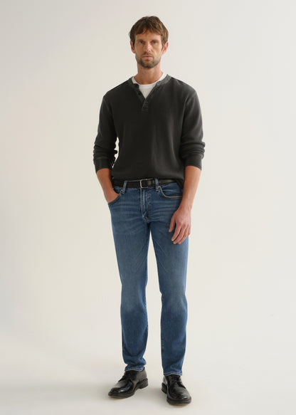 Person wearing medium blue straight leg jeans with a black long sleeve shirt and black shoes, front full body view on a light grey background.