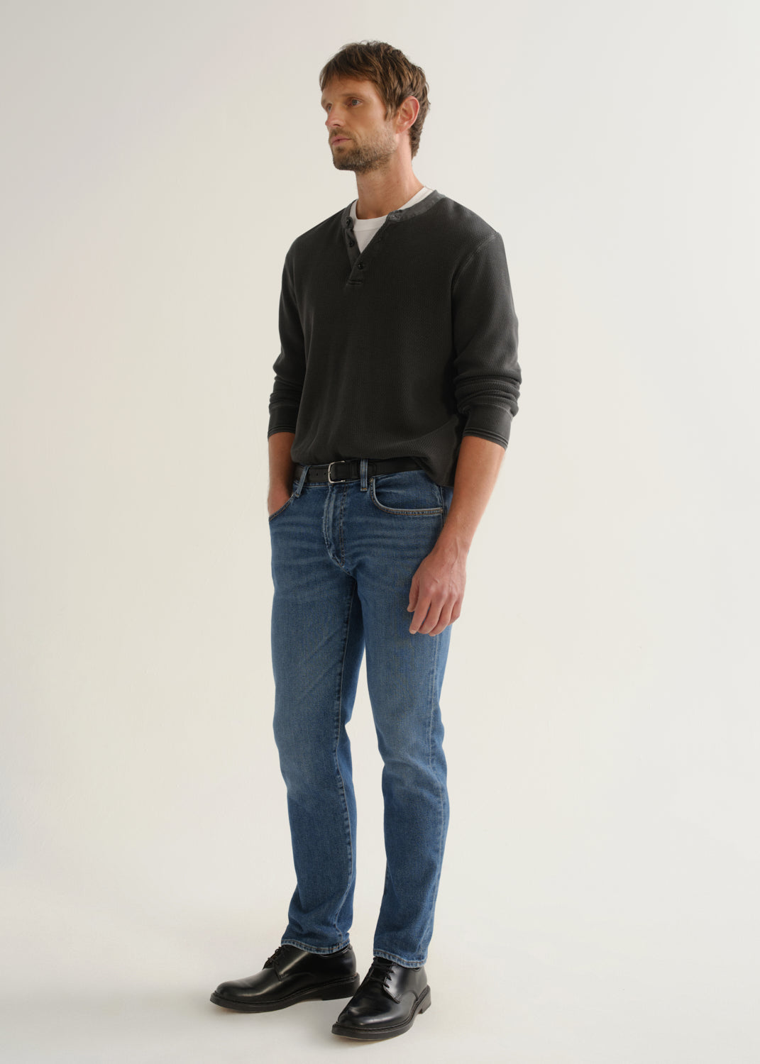 Person wearing medium blue straight leg jeans with a black long sleeve shirt and black shoes, angled front full body view on a light grey background.