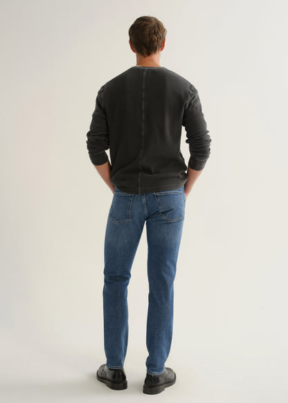 Person wearing medium blue straight leg jeans with a black long sleeve shirt and black shoes, back full body view on a light grey background.
