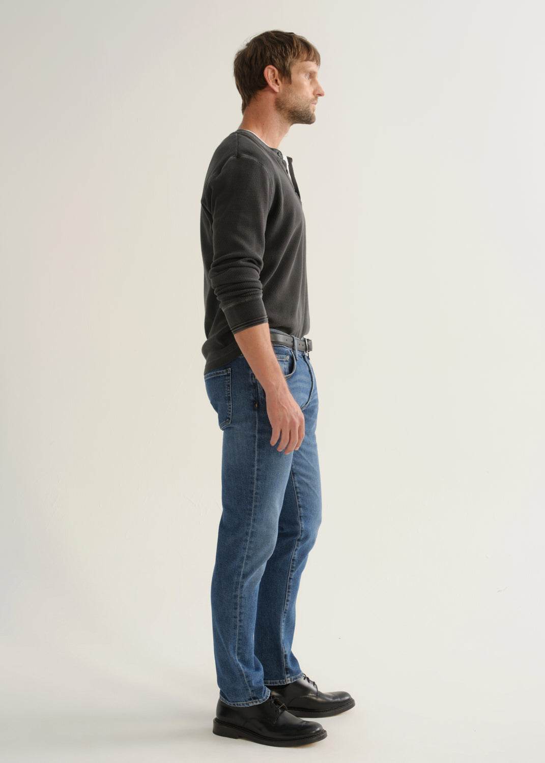 Person wearing medium blue straight leg jeans with a black long sleeve shirt and black shoes, side full body view on a light grey background.