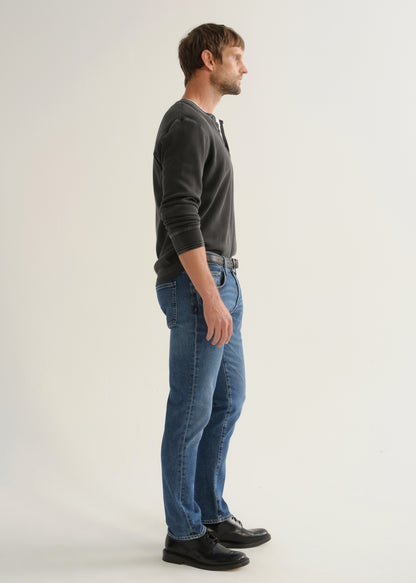 Person wearing medium blue straight leg jeans with a black long sleeve shirt and black shoes, side full body view on a light grey background.