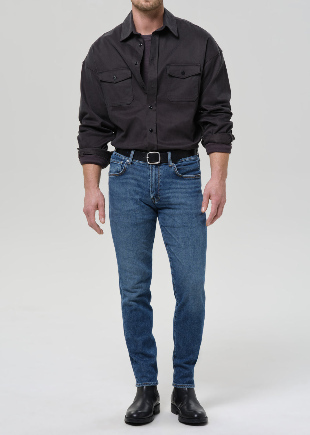 Person wearing medium blue straight leg jeans with a black button-up shirt on a grey background.
