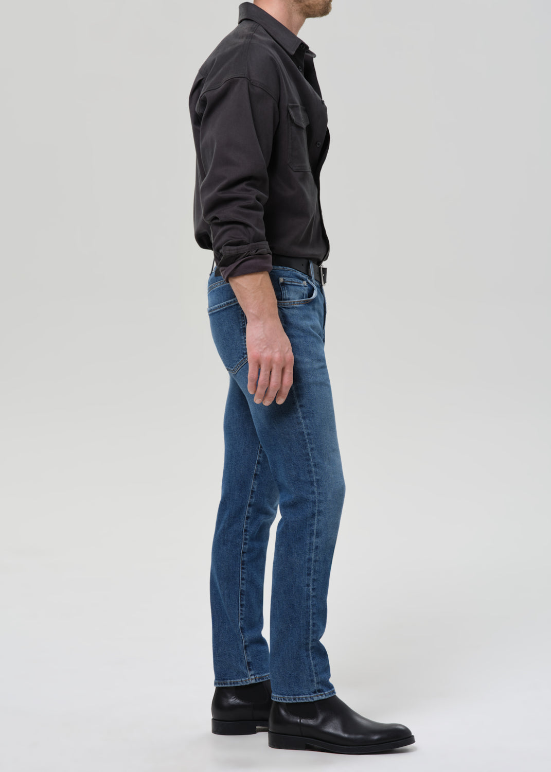 Person wearing medium blue straight leg jeans with a black button-up shirt viewed from the side on a grey background.