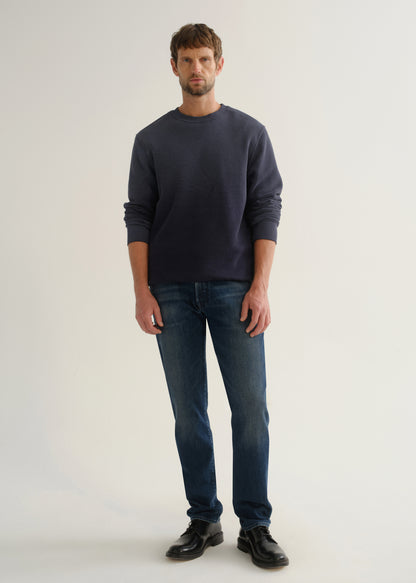 Person wearing medium dark indigo straight leg jeans with a navy long sleeve pullover and black shoes, front full body view on a light grey background.