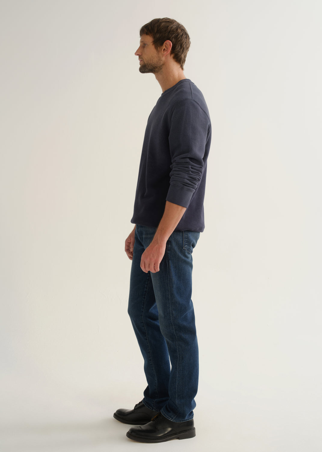 Person wearing medium dark indigo straight leg jeans with a navy long sleeve pullover and black shoes, side full body view on a light grey background.
