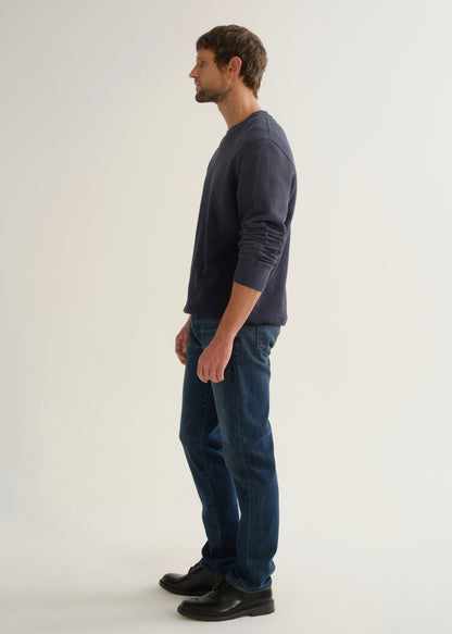 Person wearing medium dark indigo straight leg jeans with a navy long sleeve pullover and black shoes, side full body view on a light grey background.