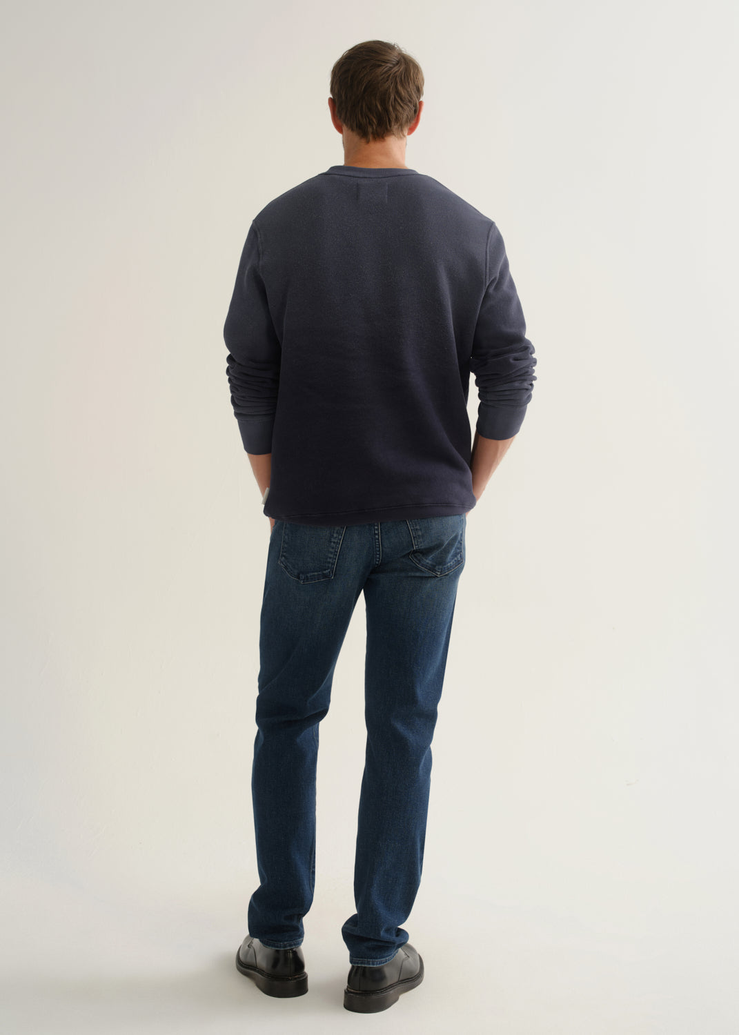Person wearing medium dark indigo straight leg jeans with a navy long sleeve pullover and black shoes, back full body view on a light grey background