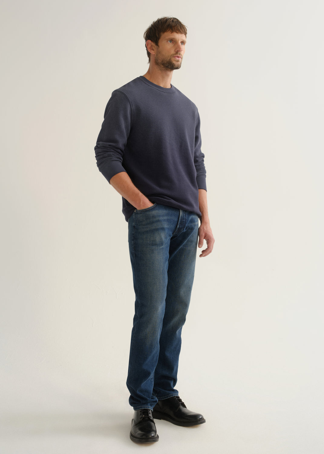 Person wearing medium dark indigo straight leg jeans with a navy long sleeve pullover and black shoes, angled front full body view on a light grey background.