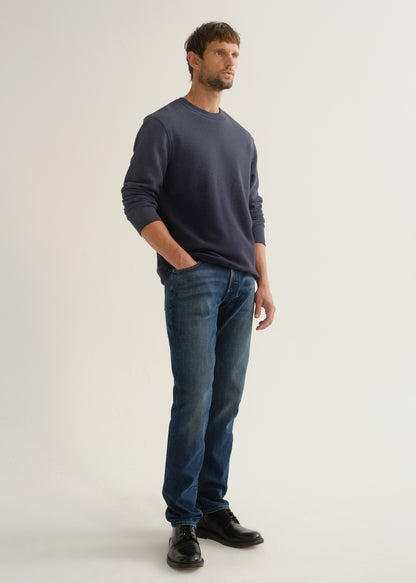 Person wearing medium dark indigo straight leg jeans with a navy long sleeve pullover and black shoes, angled front full body view on a light grey background.