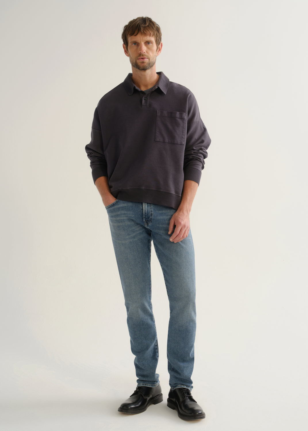 Person wearing medium blue straight leg jeans with a dark navy long sleeve collared pullover and black shoes, front full body view on a light grey background.