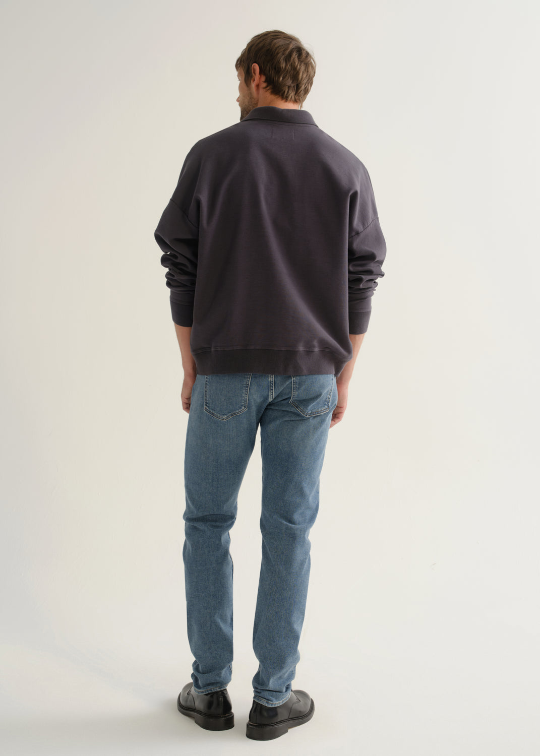 Person wearing medium blue straight leg jeans with a dark navy long sleeve collared pullover and black shoes, back full body view on a light grey background.
