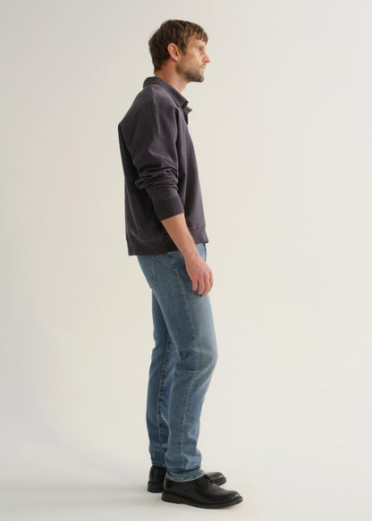 Person wearing medium blue straight leg jeans with a dark navy long sleeve collared pullover and black shoes, side full body view on a light grey background.