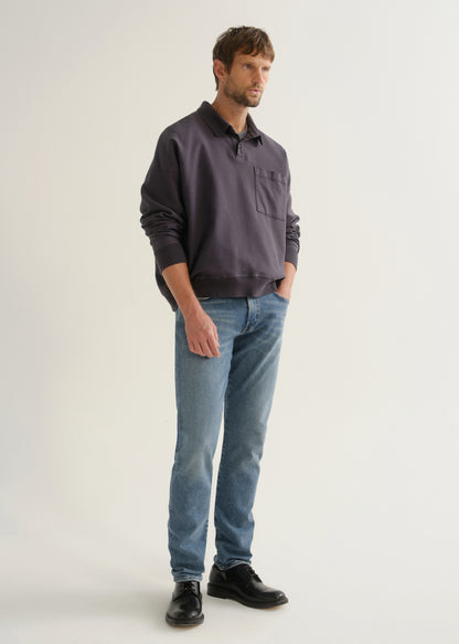 Person wearing medium blue straight leg jeans with a dark navy long sleeve collared pullover and black shoes, angled front full body view on a light grey background.
