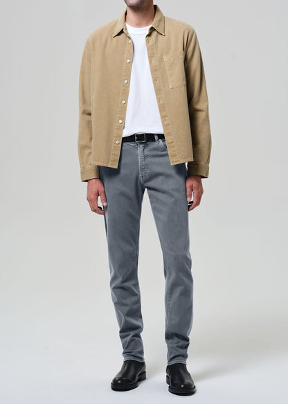 Person wearing a beige jacket, white shirt, grayish blue jeans, and black shoes on a plain background