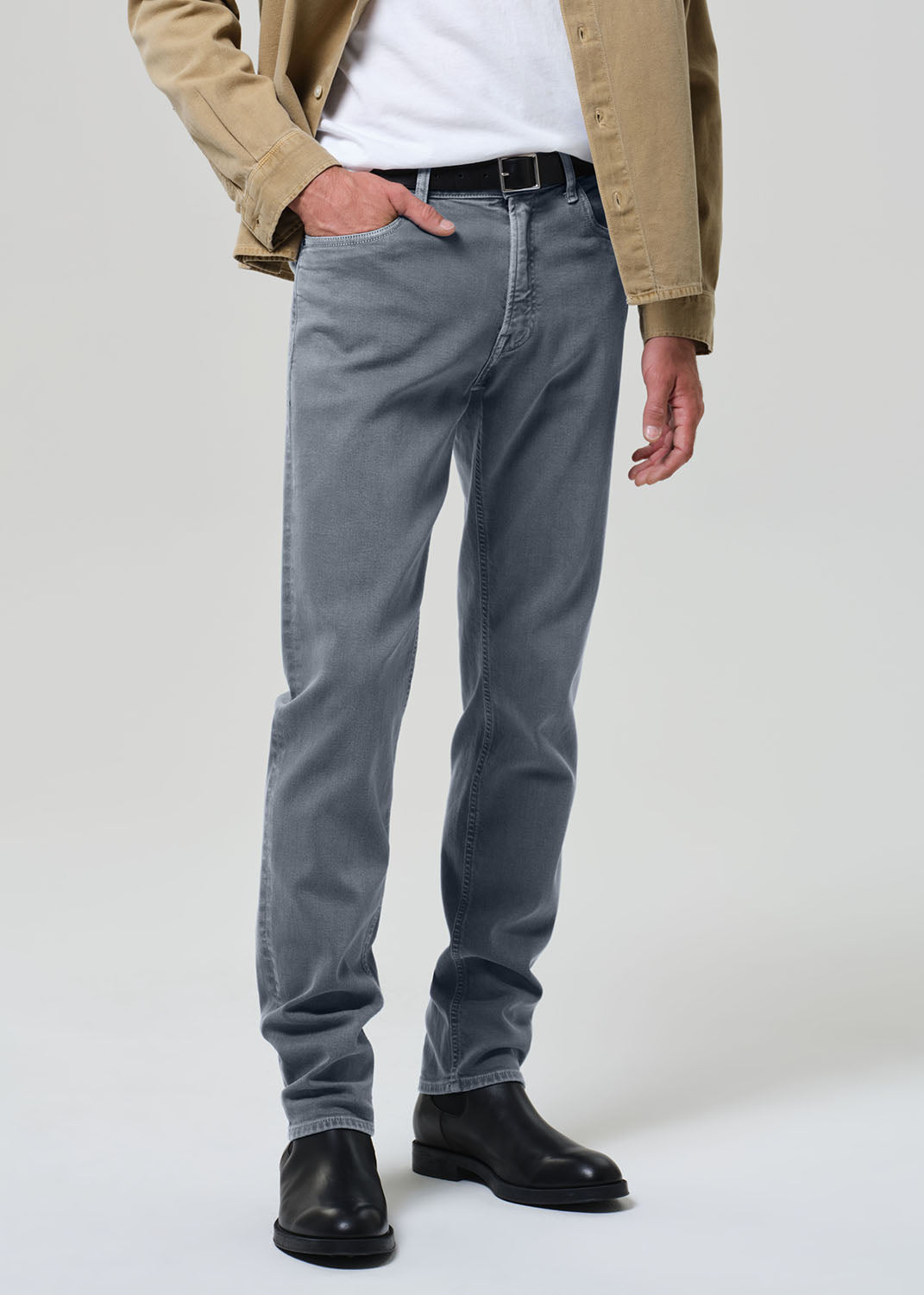 Person wearing a beige jacket, white shirt, grayish blue jeans, and black shoes on a plain background