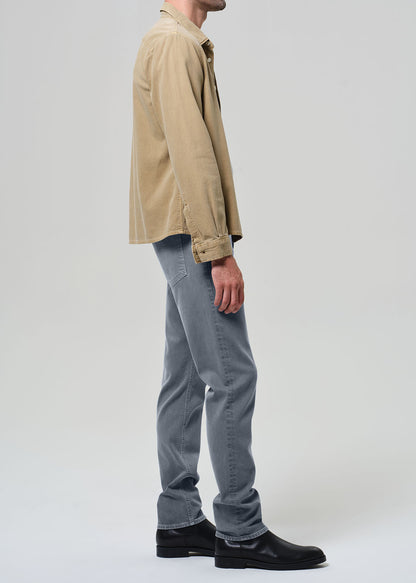 Person wearing a beige jacket, white shirt, grayish blue jeans, and black shoes on a plain background