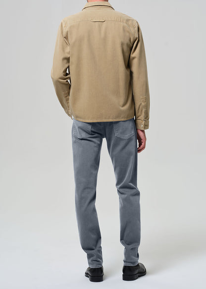 Person wearing a beige jacket, white shirt, grayish blue jeans, and black shoes on a plain background
