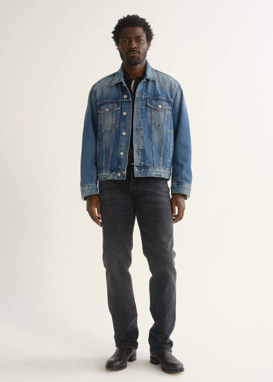 Person wearing washed black straight leg jeans with a blue denim jacket, dark shirt, and black shoes, front full body view on a light grey background.