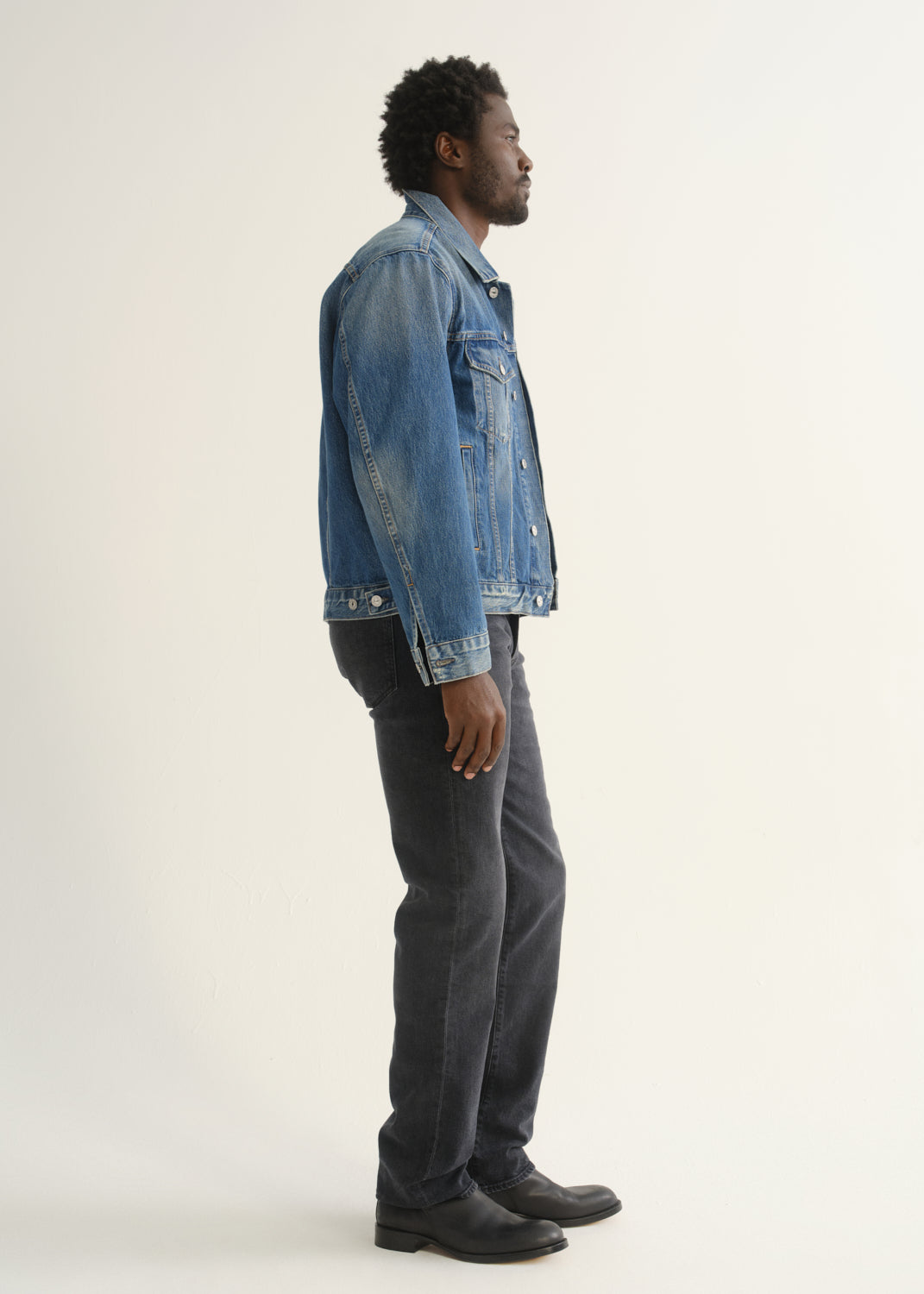 Person wearing washed black straight leg jeans with a blue denim jacket and black shoes, side full body view on a light grey background.