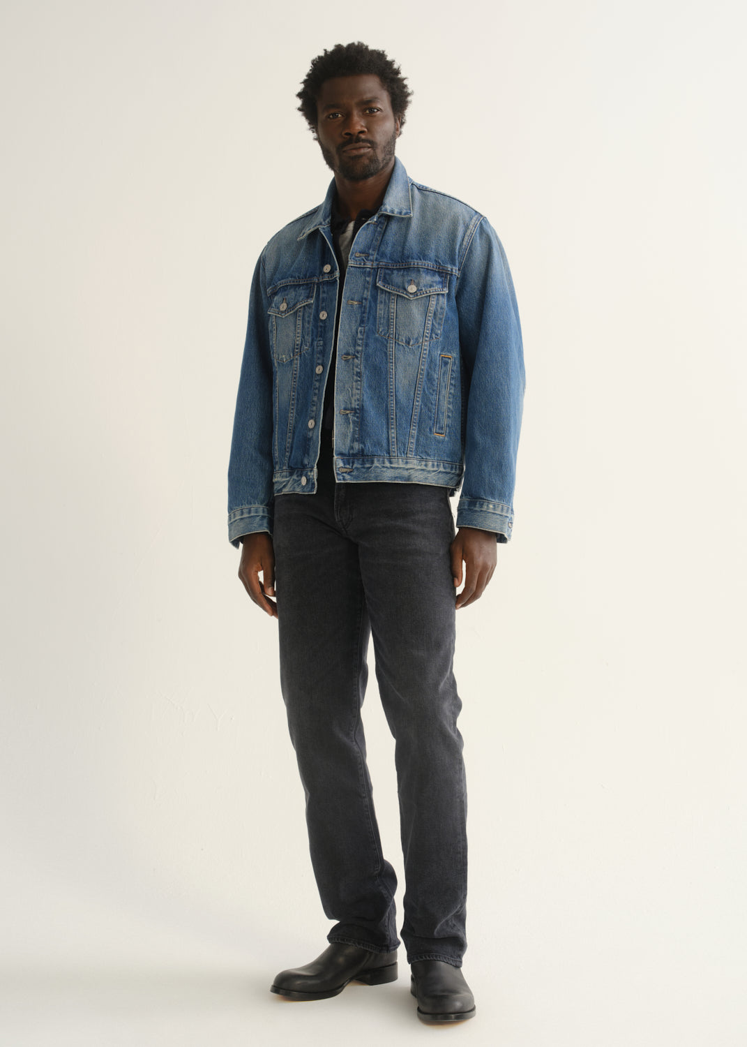 Person wearing washed black straight leg jeans with a blue denim jacket and black shoes, angled front full body view on a light grey background.