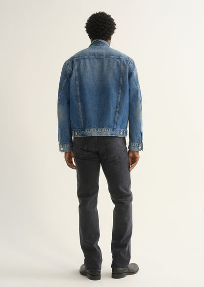 Person wearing washed black straight leg jeans with a blue denim jacket and black shoes, back full body view on a light grey background.