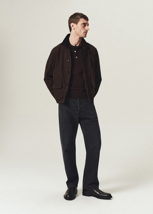 A man modeling a dark brown corduroy jacket with a black collar, worn over a dark brown sweater, dark gray jeans, and black leather shoes, against a light gray studio background.
