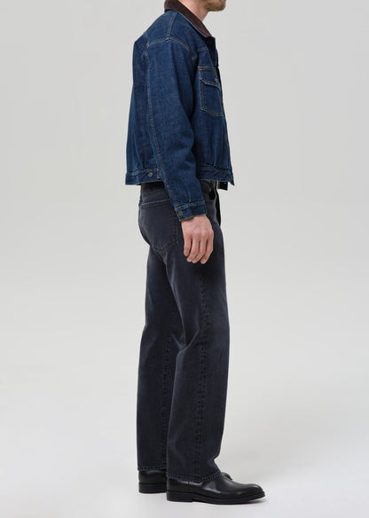 Person wearing a blue denim jacket and dark jeans on a grey background