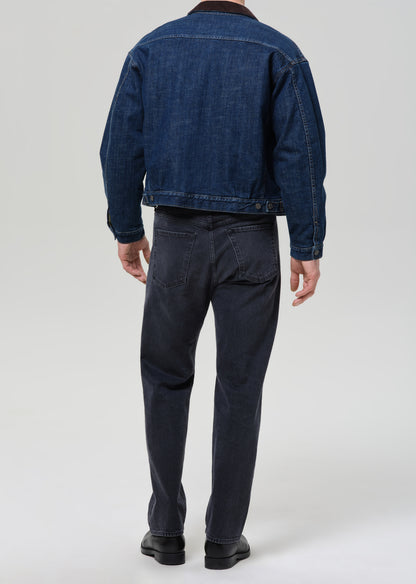 Person wearing a blue denim jacket and dark pants on a grey background