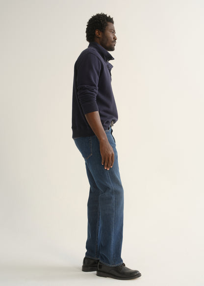 Person wearing medium dark indigo straight leg jeans with a navy long sleeve collared pullover and black shoes, side full body view on a light grey background.