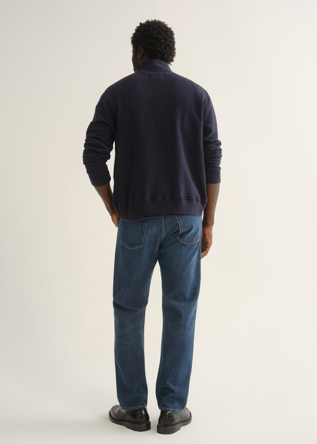 Person wearing medium dark indigo straight leg jeans with a navy long sleeve collared pullover and black shoes, back full body view on a light grey background.