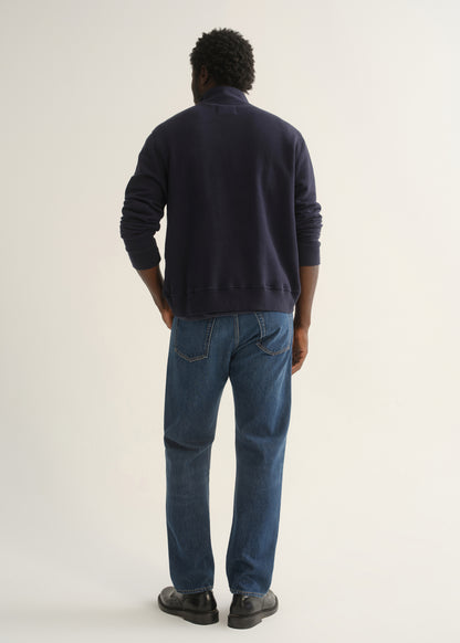 Person wearing medium dark indigo straight leg jeans with a navy long sleeve collared pullover and black shoes, back full body view on a light grey background.