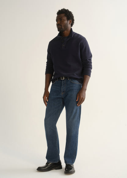 Person wearing medium dark indigo straight leg jeans with a navy long sleeve collared pullover and black shoes, angled front full body view on a light grey background.