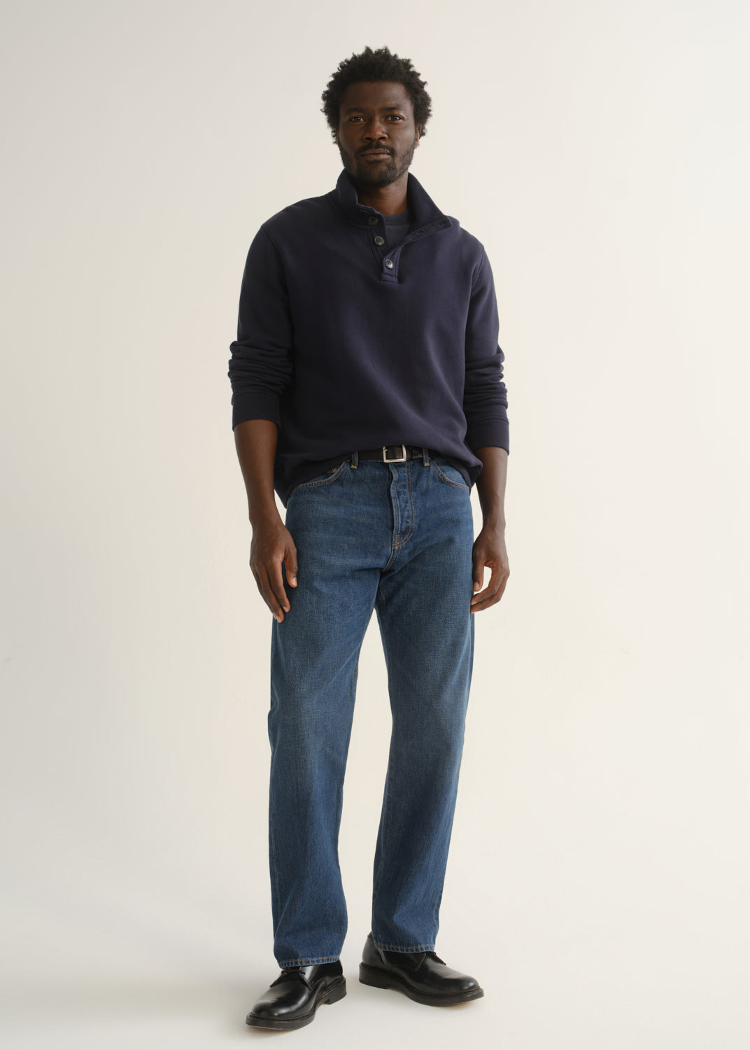 Person wearing medium dark indigo straight leg jeans with a navy long sleeve collared pullover and black shoes, front full body view on a light grey background.