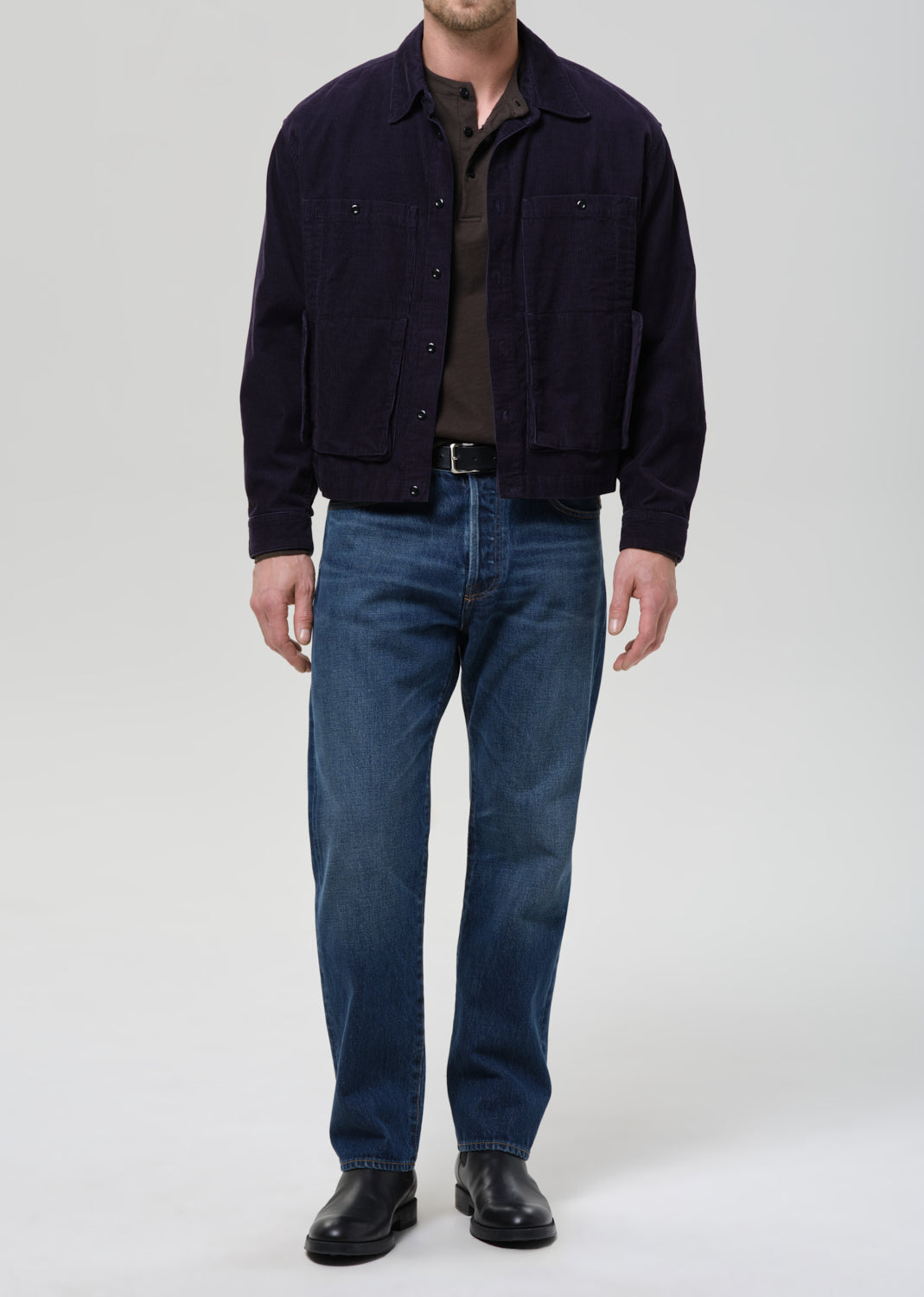 A man wearing a dark navy corduroy jacket over a dark brown shirt, blue jeans, and black leather boots, standing against a grey background.