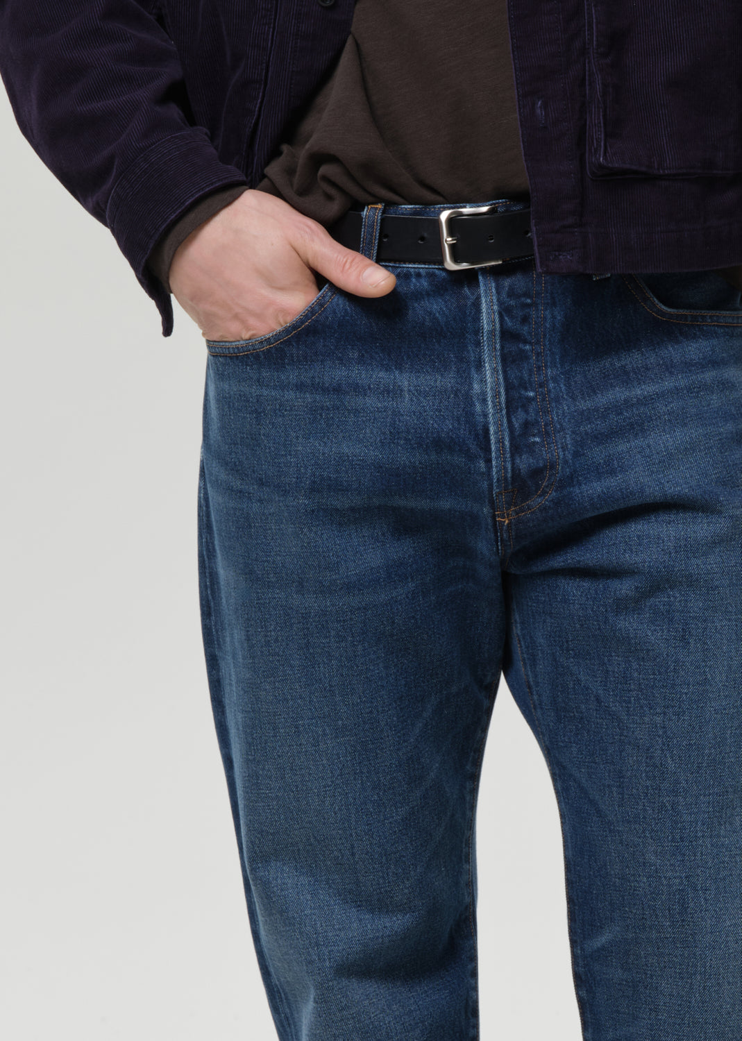 Close-up of a person wearing dark blue denim jeans with a black belt, a dark brown shirt, and a dark navy jacket, set against a grey background.
