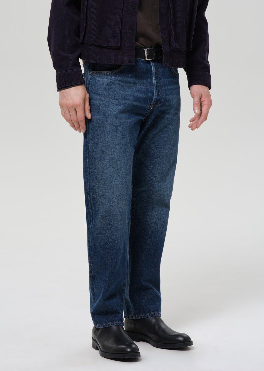A person wearing dark blue straight-leg denim jeans, a dark navy jacket, a dark brown shirt, and black leather boots, photographed against a grey background.