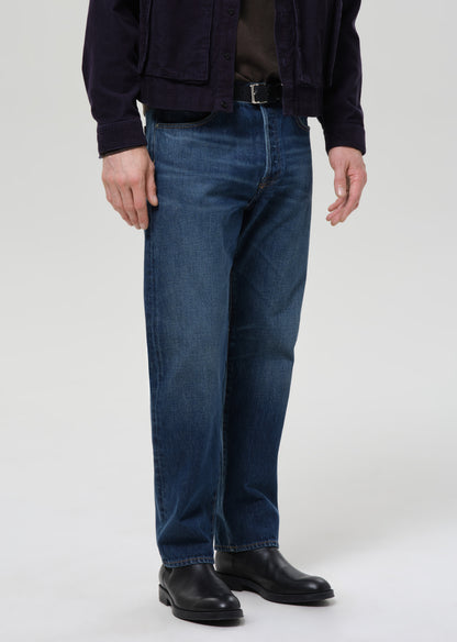 A person wearing dark blue straight-leg denim jeans, a dark navy jacket, a dark brown shirt, and black leather boots, photographed against a grey background.