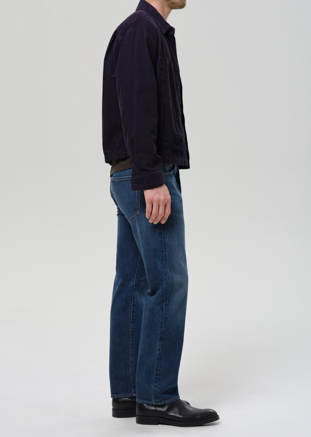 A side-profile view of a person wearing a dark navy corduroy jacket, blue jeans, and black leather shoes against a grey background.