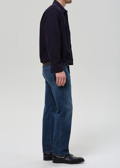 A side-profile view of a person wearing a dark navy corduroy jacket, blue jeans, and black leather shoes against a grey background.