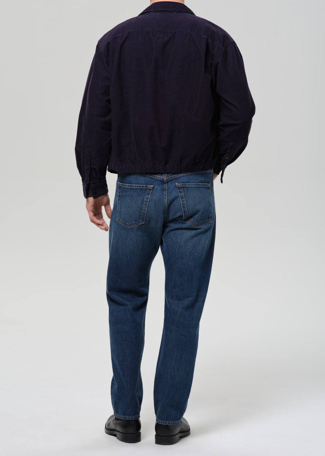 Back view of a person wearing a dark navy jacket and blue jeans against a grey background.