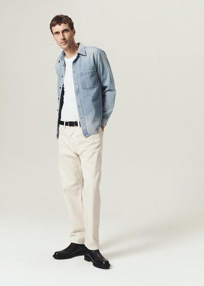 A man modeling a light blue denim shirt jacket over a white t-shirt, paired with cream-colored trousers and black leather shoes, against a plain light gray studio background.