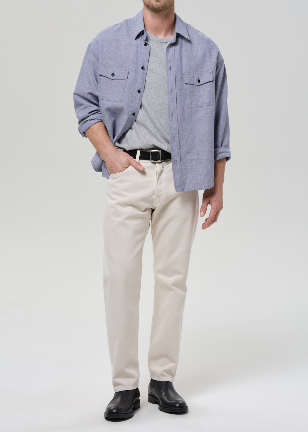 Man wearing a blue checkered shirt, grey t-shirt, beige pants, and black shoes on a grey background