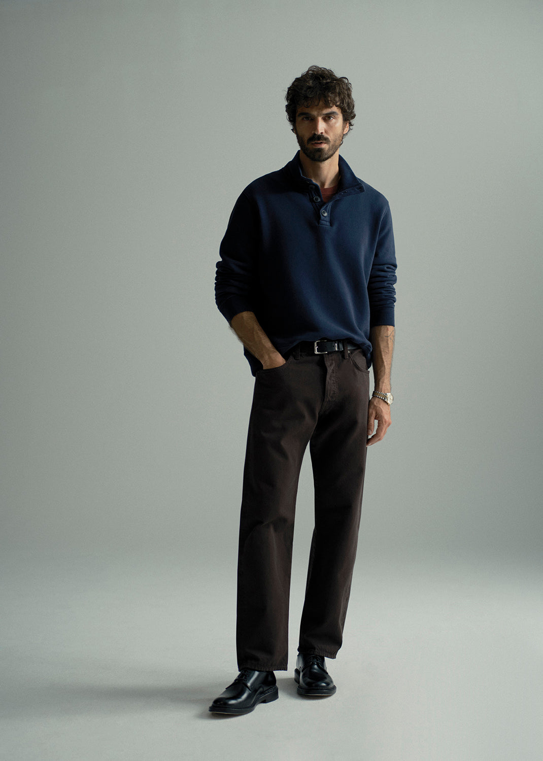 A man modeling a dark navy long-sleeve polo shirt paired with dark brown trousers and black leather dress shoes, standing against a neutral light gray studio background.