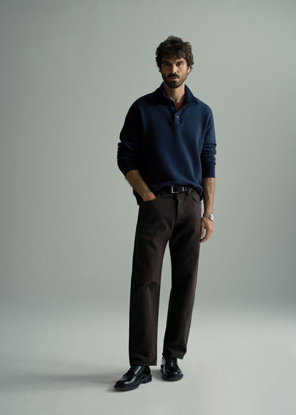 A man modeling a dark navy long-sleeve polo shirt paired with dark brown trousers and black leather dress shoes, standing against a neutral light gray studio background.
