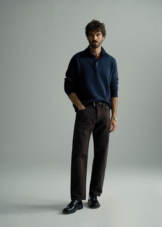 A man modeling a dark navy long-sleeve polo shirt paired with dark brown trousers and black leather dress shoes, standing against a neutral light gray studio background.