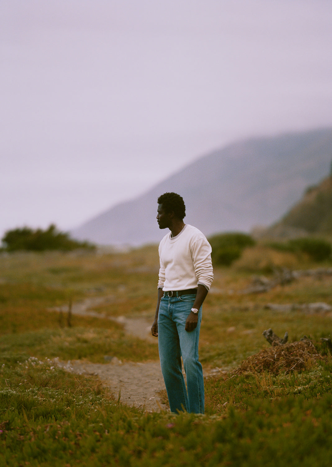 A man stands on a dirt path in an open grassy landscape, wearing a cream long-sleeve knit top and blue jeans, with hazy mountains in the background.