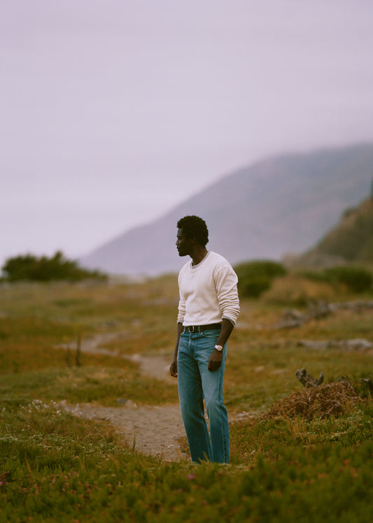 A man stands on a dirt path in an open grassy landscape, wearing a cream long-sleeve knit top and blue jeans, with hazy mountains in the background.