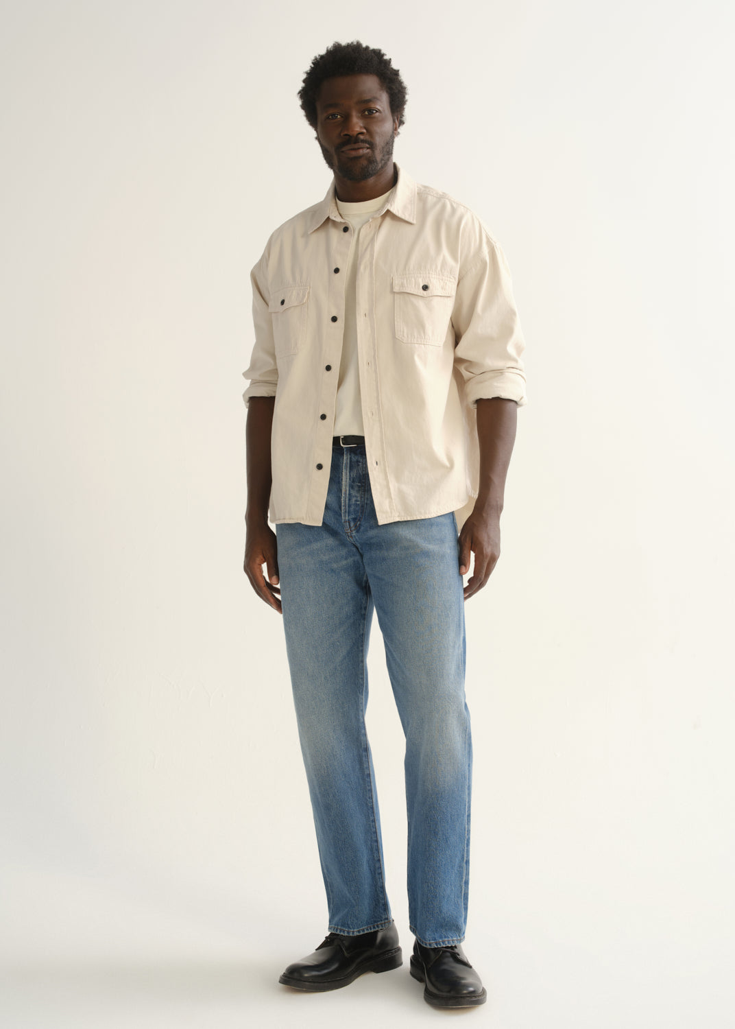 Person wearing light blue straight leg jeans with a cream button-up shirt and black shoes, front full body view on a light grey background.